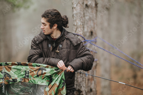 Canvas Print A young man adjusts a camouflage tarp while preparing a campsite in a mountain forest