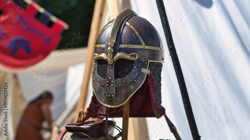 A detailed, reconstructed Viking helmet or Anglo-Saxon spangenhelm with nasal guard and chain mail, displayed at a historical camp during a medieval fair.