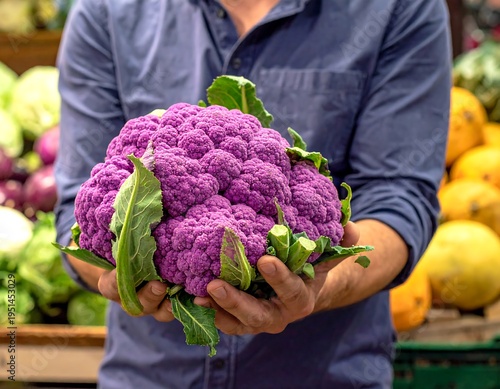 Man holding vibrant purple cauliflower, surrounded by produce in a market, wearing a blue shirt