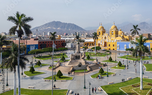 High-resolution aerial drone view of the Plaza de Armas de Trujillo in Trujillo, featuring colonial architecture, colorful buildings, and the central monument surrounded by palm trees.