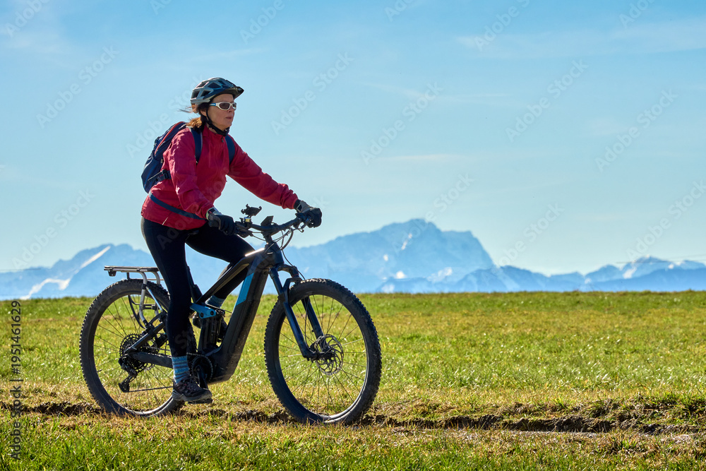 Obraz premium Middle aged woman cycling on an electric bike across green spring meadow in Upper Bavaria with alpine mountain range and Zugspitze silhouette in background under clear blue sky.
