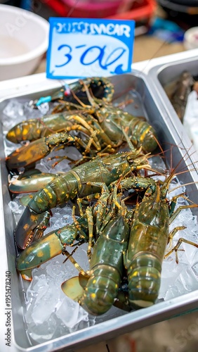 Green lobsters piled on crushed ice in a metallic tray under a price sign, outdoor food market stall