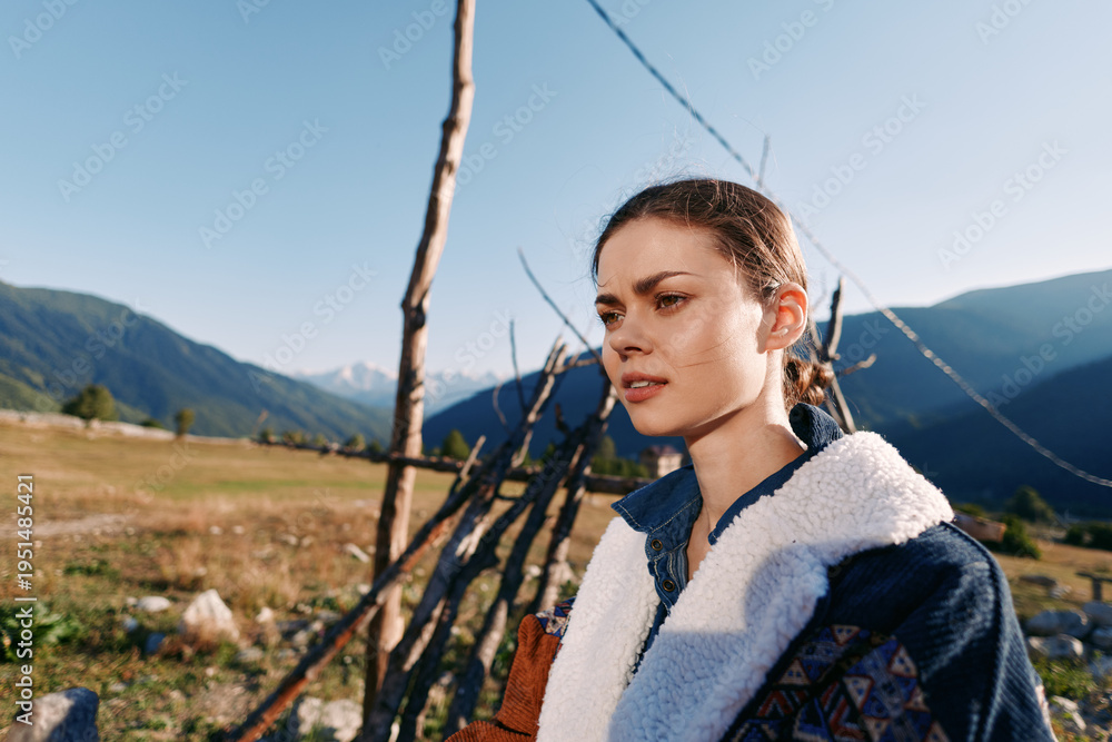 Naklejka premium Woman portrait in countryside by a wooden fence with mountains in the background, outdoor rural scene at golden hour. Young female looking thoughtful amid nature and sunlight