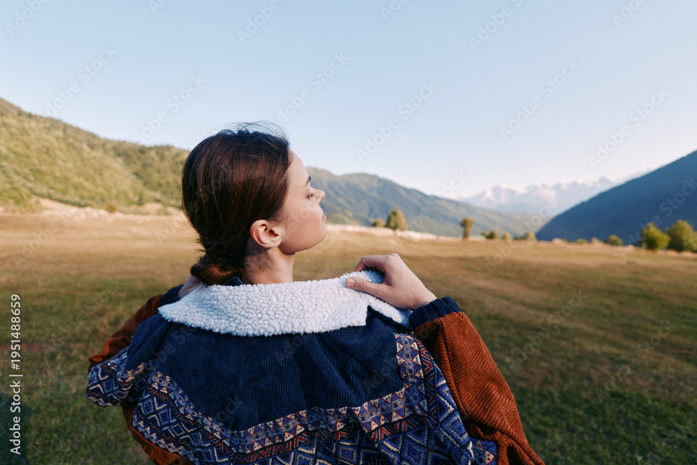 Naklejka premium Woman in patterned jacket standing in meadow outdoors, back portrait gazing at mountain range, autumn light, serene landscape, nature travel and contemplative moment.