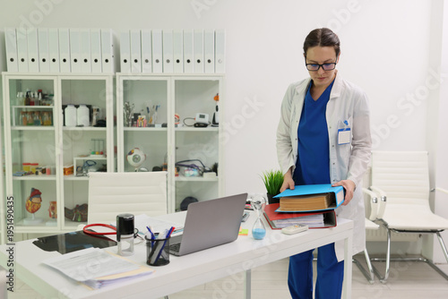 In a bright medical office, a focused doctor in a white coat sorts through colorful patient files. The workspace features a laptop, plants, and organized supplies, highlighting dedication.