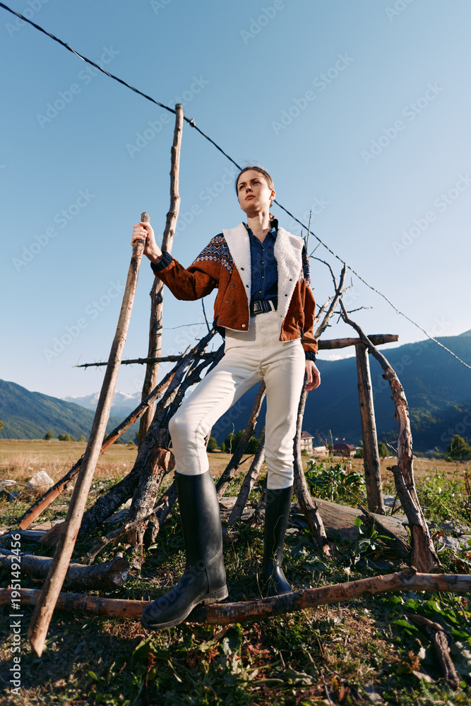 Obraz premium Woman in rubber boots holds a wooden stick standing by a rustic fence in countryside, wearing patterned jacket and white trousers, posing confidently against mountains and clear sky.