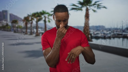 Man pinching nose and waving hand at seaside marina promenade with boats and palm trees visible; disgust.