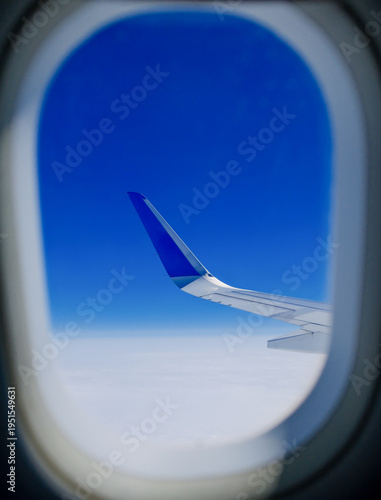 Airplane wing and white clouds seen through oval passenger window porthole during daytime flight travel