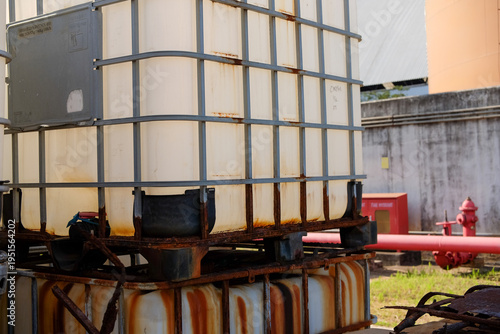 Industrial Intermediate Bulk Containers Stacked in an Outdoor Facility Showcasing Prominent Rust and Wear on Metal Cages Surrounded by a Concrete Wall and Red Pipes