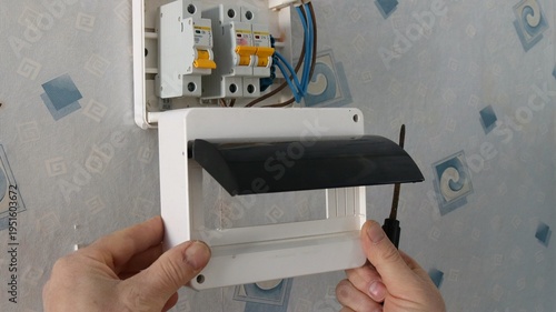 A man's hands hold a screwdriver, securing the plastic casing of a household electricity meter hanging on a wall covered in old wallpaper; the man is repairing and installing a home electricity meter.