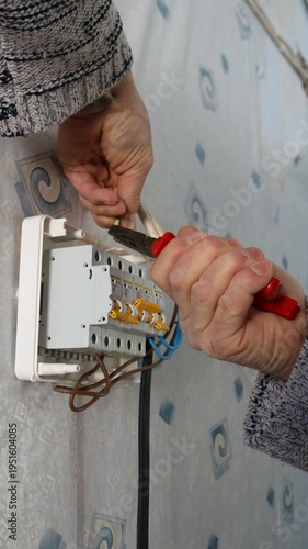 A man's hands hold pliers and secure the wires going to a wall-mounted home electrical distribution box with circuit breakers. DIY wall-mounted electrical equipment installation using a hand tool.