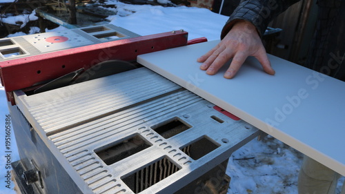 carpenter's hand presses a laminated board against the surface of an operating circular saw table to cut off a section—processing construction material on a miter saw in an outdoor carpentry workshop
