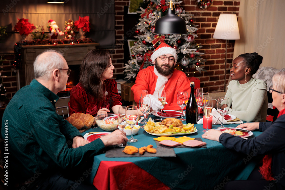 Obraz premium Man in santa claus costume sitting at table with diverse people, enjoying festive dinner on christmas eve festivity. Young person in suit spreading positivity during december holidays.