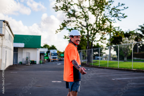 Worker in orange shirt and helmet smiles while standing in a busy outdoor work site in New Zealand