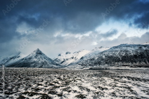 A wide-angle landscape featuring jagged snow-covered mountains and a frozen tundra under a heavy, dramatic winter sky. Ideal for travel and nature themes.
