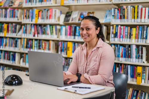 A woman sits at a desk in a library typing on a laptop while smiling
