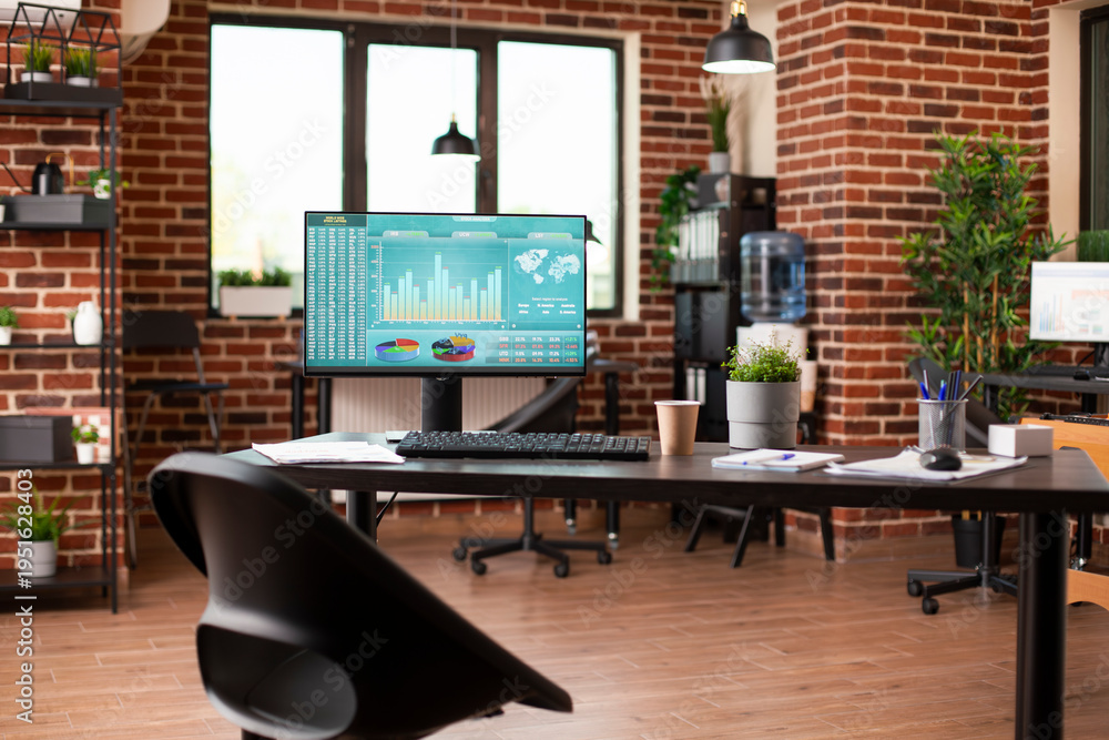 Fototapeta premium Empty startup office having modern desk with computer monitor displaying financial charts. Desktop pc, shelves with folders, boxes and plant decorations completing well organized brick wall workspace.