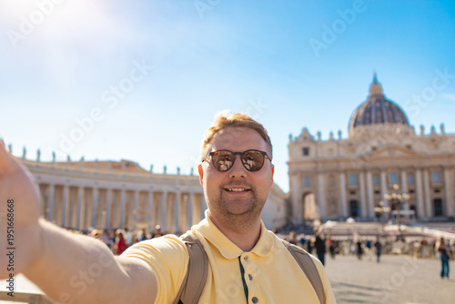 Smiling male tourist taking selfie in St Peter Square with St Peter Basilica in background on sunny day.