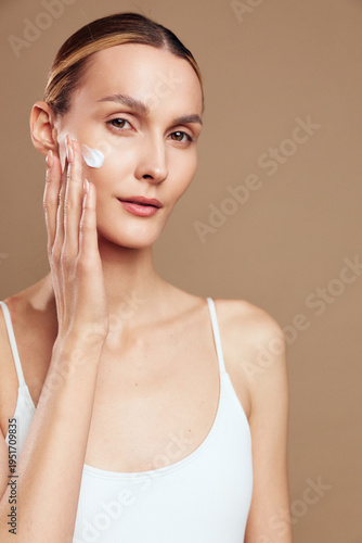 Foto A young woman with clear skin applies white cream to her face while looking at the camera