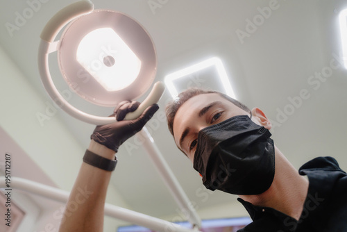 The dentist looks at the camera and shines a lamp. Doctor's face in close-up. A dentist in a black uniform at a dental clinic. A doctor wearing a black mask. The dentist's focused gaze. 