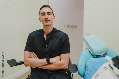 A young smiling dentist at his workplace. Specialist in oral cavity treatment. A doctor in a black uniform. A doctor with a watch on his right hand. Interior of a dental office.