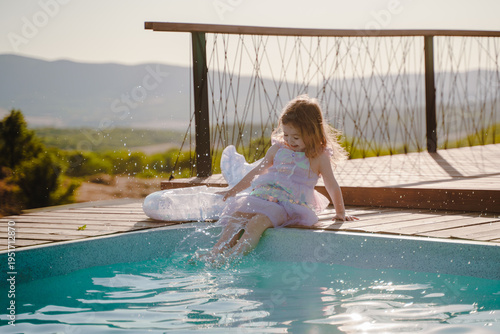 A happy and contented little girl is sitting in a mermaid dress and splashing her legs in the pool. A child's rest at sunset.