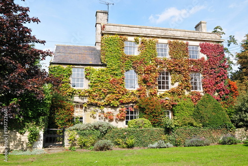 An elegant stone manor house adorned with vibrant autumn ivy in shades of red and green. This classic architectural scene, set behind a manicured hedge and garden lawn, represents luxury real estate.