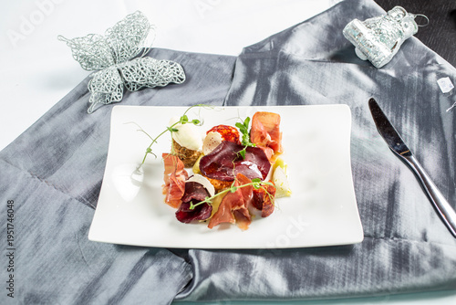 Modernly arranged appetizer with thinly sliced ​​prosciutto, cheese and side dishes, decorated with fresh sprouts on a white plate.