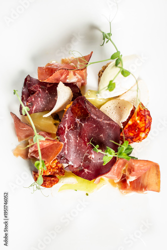 Modernly arranged appetizer with thinly sliced ​​prosciutto, cheese and side dishes, decorated with fresh sprouts on a white plate.