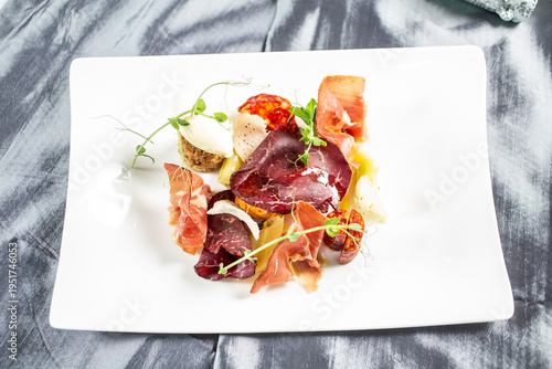 Modernly arranged appetizer with thinly sliced ​​prosciutto, cheese and side dishes, decorated with fresh sprouts on a white plate.