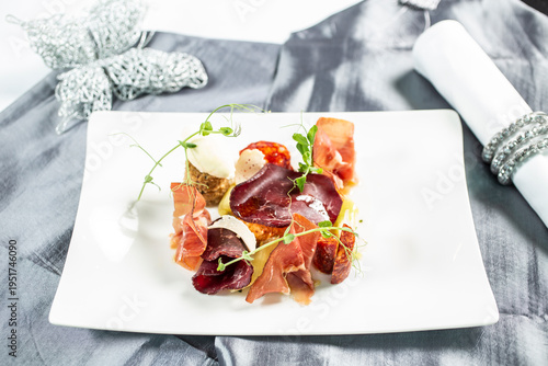 Modernly arranged appetizer with thinly sliced ​​prosciutto, cheese and side dishes, decorated with fresh sprouts on a white plate.