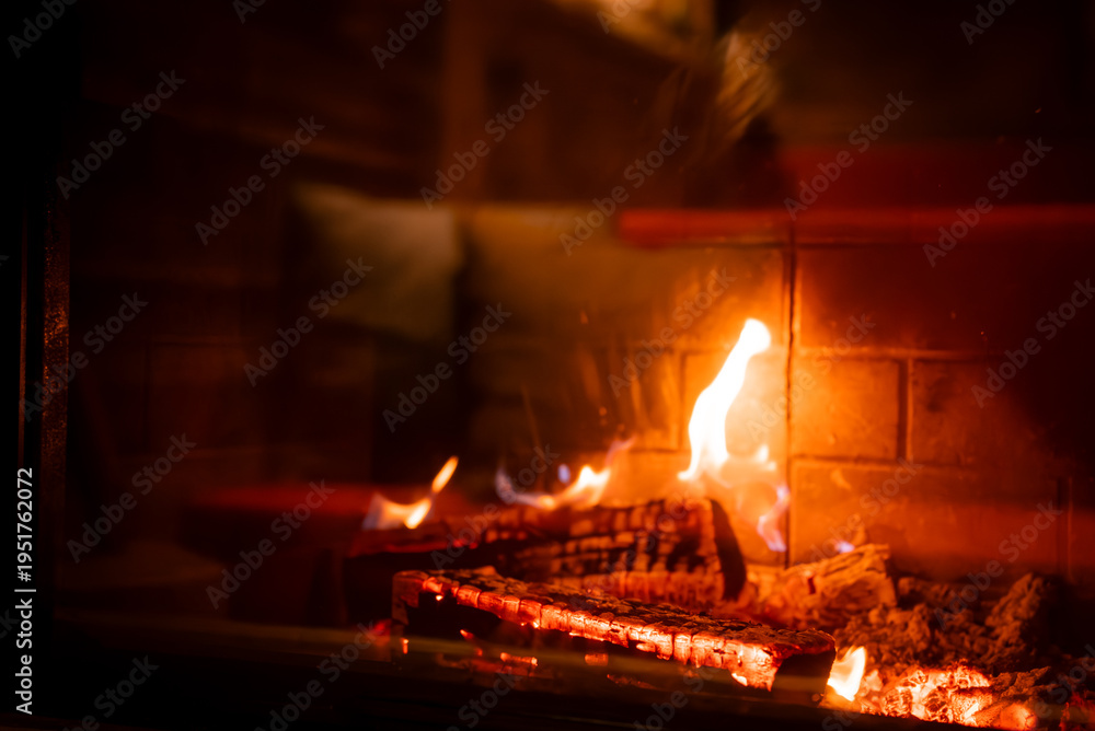 Fototapeta premium Charred logs and embers glow in a brick lined fireplace at a San Domenico lodge in the Italian Alps, with warm wood interiors and soft cushions visible beyond.