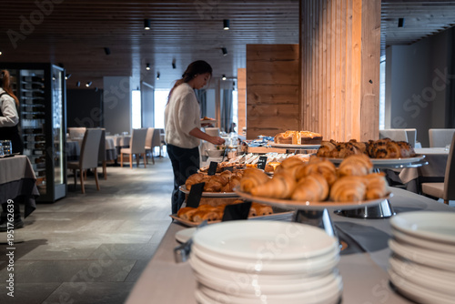 A modern dining room in the San Domenico ski resort, Italian Alps, shows a breakfast buffet with golden croissants and pastries as a guest serves herself, morning mood.