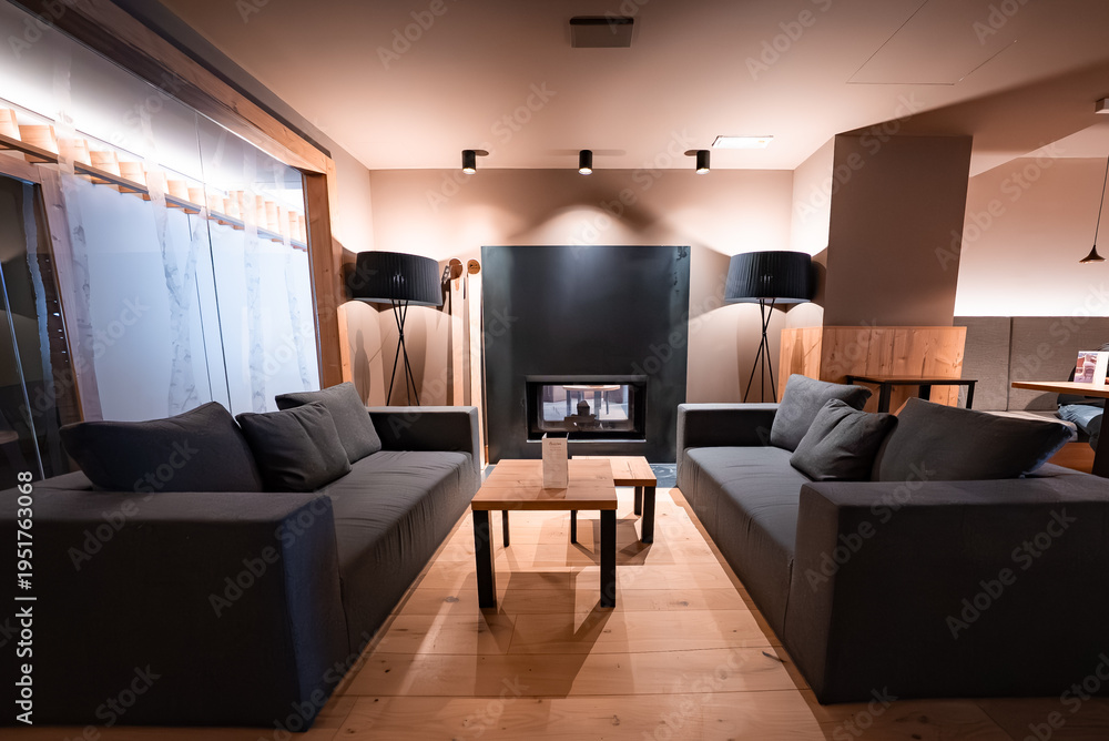 Fototapeta premium Two charcoal gray sofas face wooden tables by a built in fireplace in San Domenico, Italian Alps. Tripod lamps and recessed lights warm the rustic wood interior.
