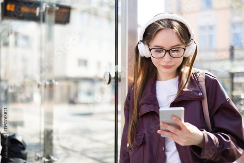 Student learning online at bus stop in city. Distance learning with smartphone. Girl with headphones outdoors. Young woman waiting for public transport, tram. Technology lifestyle, urban commute