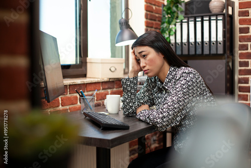 Fotografija Tired businesswoman sitting at desk, face in hand, struggling to focus after an extended workday in modern workplace