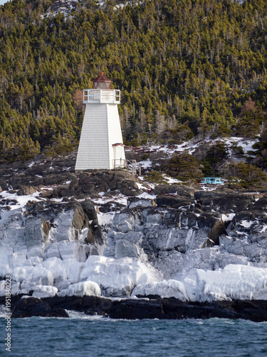 A white light tower stands along the frozen winter coast, surrounded by ice‑covered rocks and a backdrop of dark evergreen trees, creating a stark and atmospheric maritime scene.