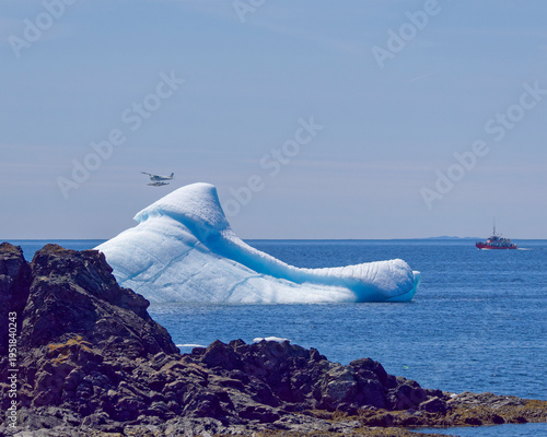 A towering iceberg rises behind large foreground rocks as a small plane passes overhead, while a distant tour boat moves quietly across the background, creating a dramatic and dynamic polar scene.