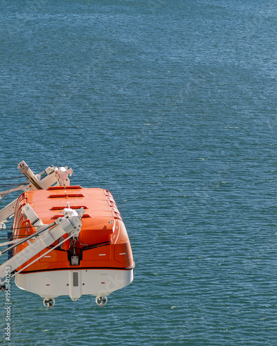 An enclosed lifeboat with an orange top and white hull attached to a quick release system, its bright safety colors standing out sharply against the surrounding marine environment.