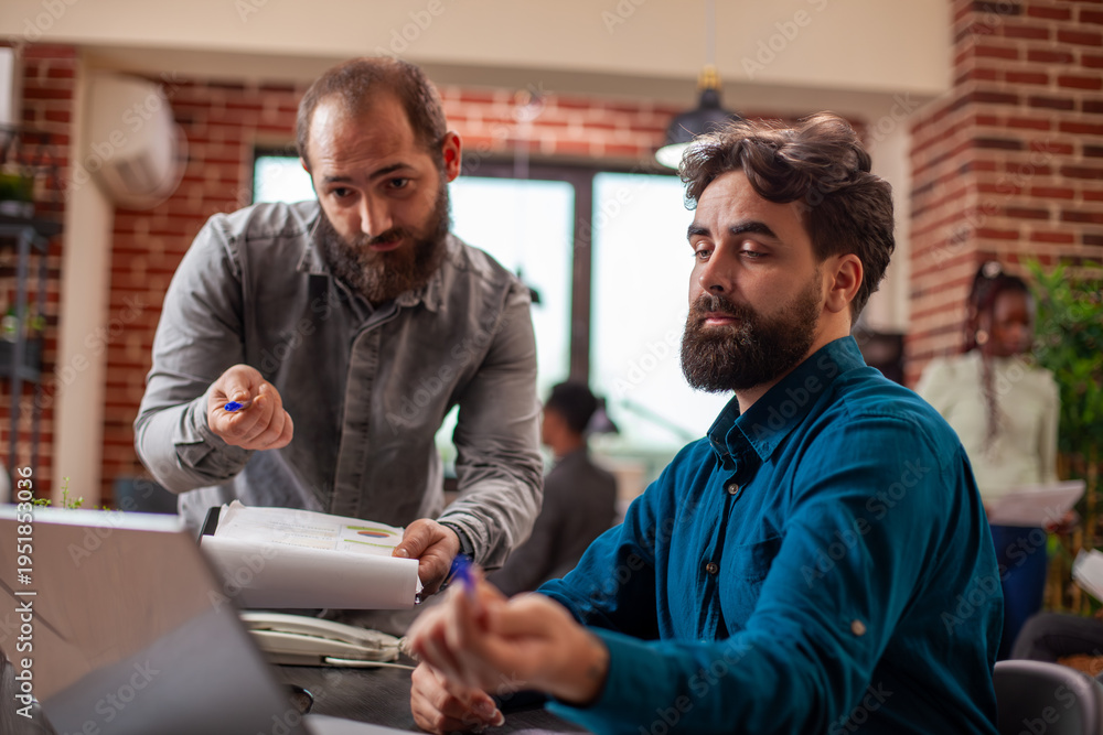 Fototapeta premium Caucasian male colleagues, one with clipboard, are working together at office desk. Businessmen point at laptop screen, reviewing documents and planning a new marketing strategy for startup company.