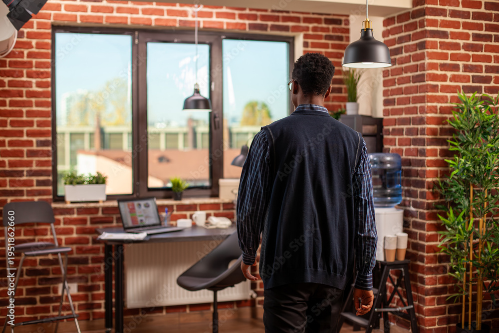Fototapeta premium Black businessman walking into brick wall room, heading to sit at his well organized office desk. African american male manager entering modern workspace, ready for work at startup company.
