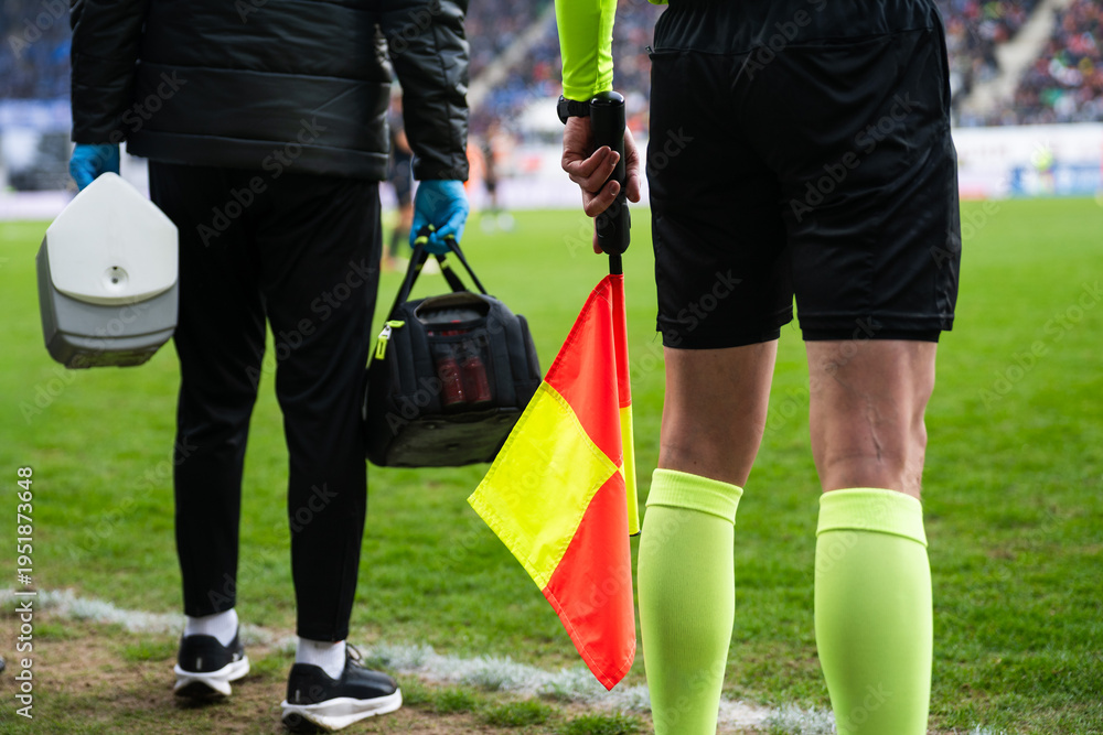 Obraz premium Close-up of an assistant referee holding a yellow-red flag beside a medical staff member at a professional soccer stadium sideline.