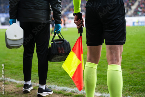 Close-up of an assistant referee holding a yellow-red flag beside a medical staff member at a professional soccer stadium sideline.