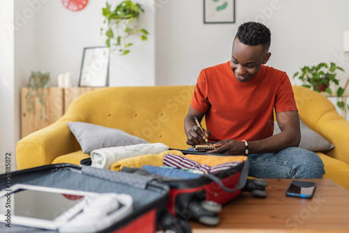 Tableau sur toile Young African man sitting on sofa preparing suitcase for travel, making checklis