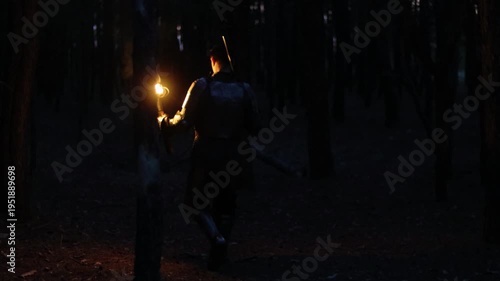 Viewed from behind, a knight in full armor, holding a sword and torch, walks through a dark night forest. A medieval warrior in the dark.