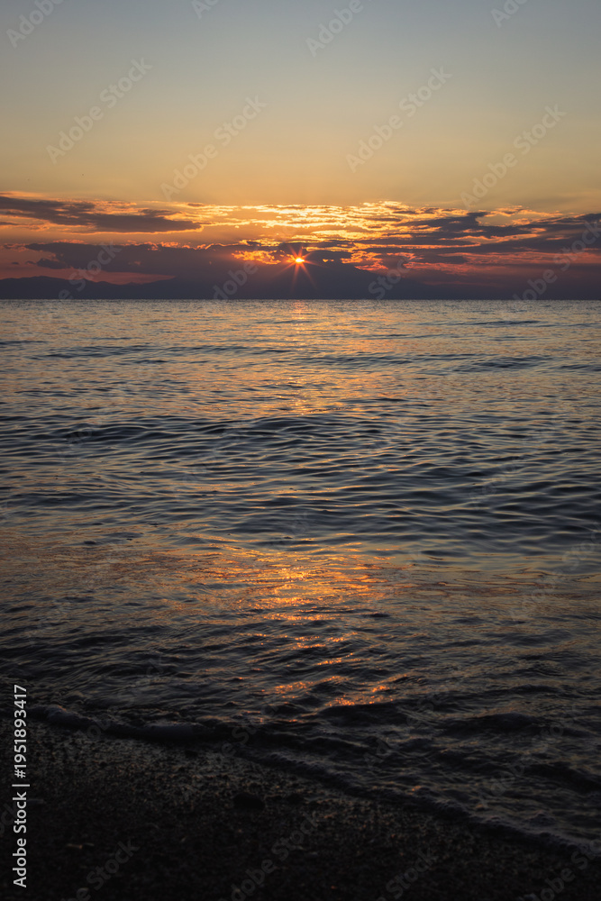 Obraz premium Serene sunset scene with sun breaking through dramatic cloud layers over tranquil ocean water. Golden and orange light reflects on gentle waves, perfect for travel, meditation, and nature concepts.