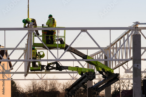 construction workers in high-visibility gear on an aerial lift installing steel roof truss structure against a clear sky