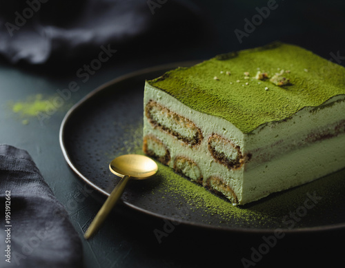 A layered matcha tiramisu cake, elegantly dusted with green tea powder, is presented on a dark plate alongside a gold spoon. This sophisticated dessert delivers rich flavors and an exquisite visual ap
