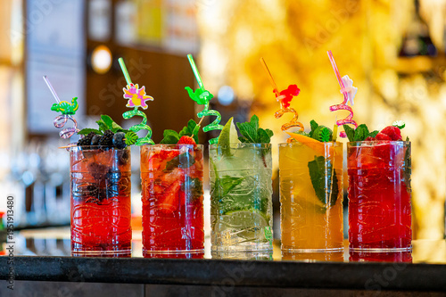 Colorful drinks sit on the bar counter. Six different drinks are arranged on the bar counter with fruit and decorative straws.