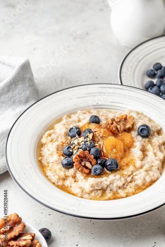 Oatmeal porridge with caramelized bananas, nuts, blueberries and honey in a plate on light gray background. Healthy vegan dish or morning breakfast concept